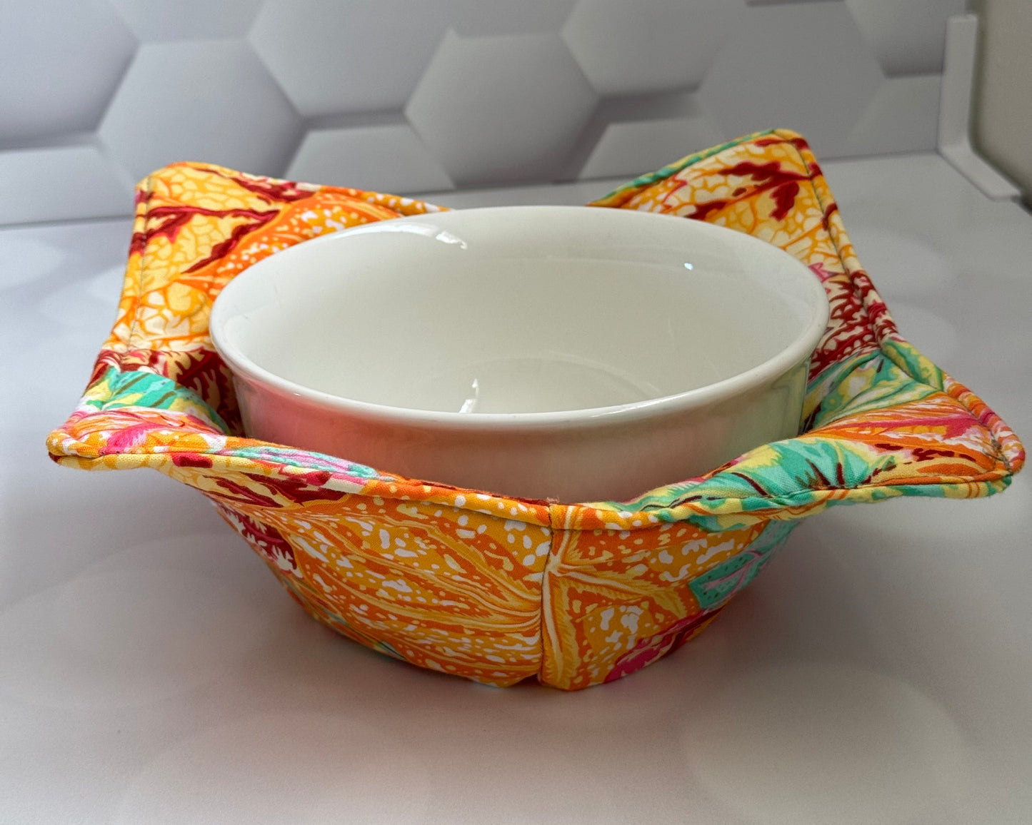 A colorful orange leaf-shaped bowl cozy placed on a white surface with a hexagonal background.